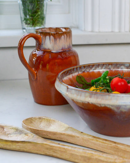 Ceramic pitcher and bowl with tomato-based dish on a white surface with wooden salad servers