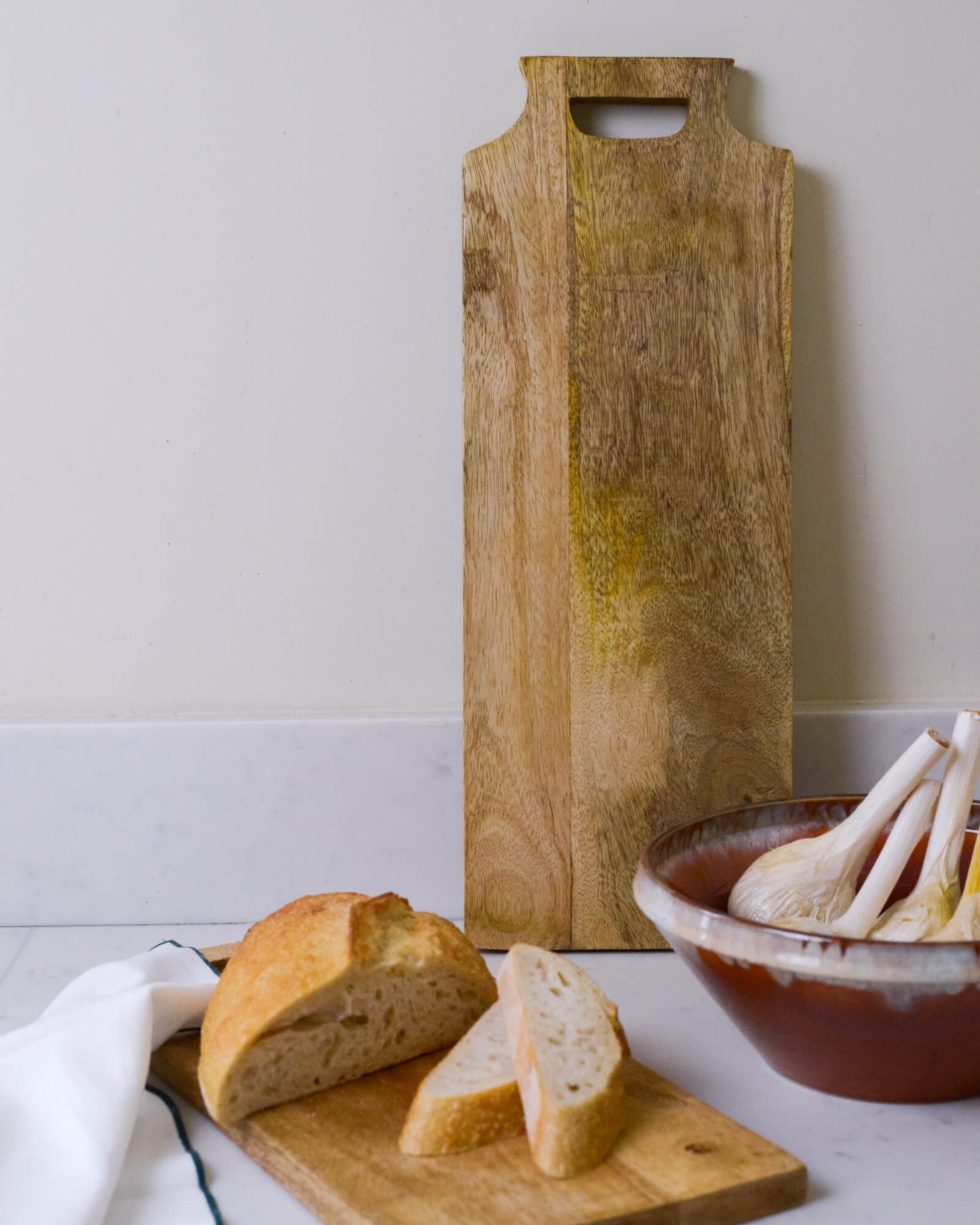 Wooden cutting board with bread and a ceramic bowl on a white marble surface.