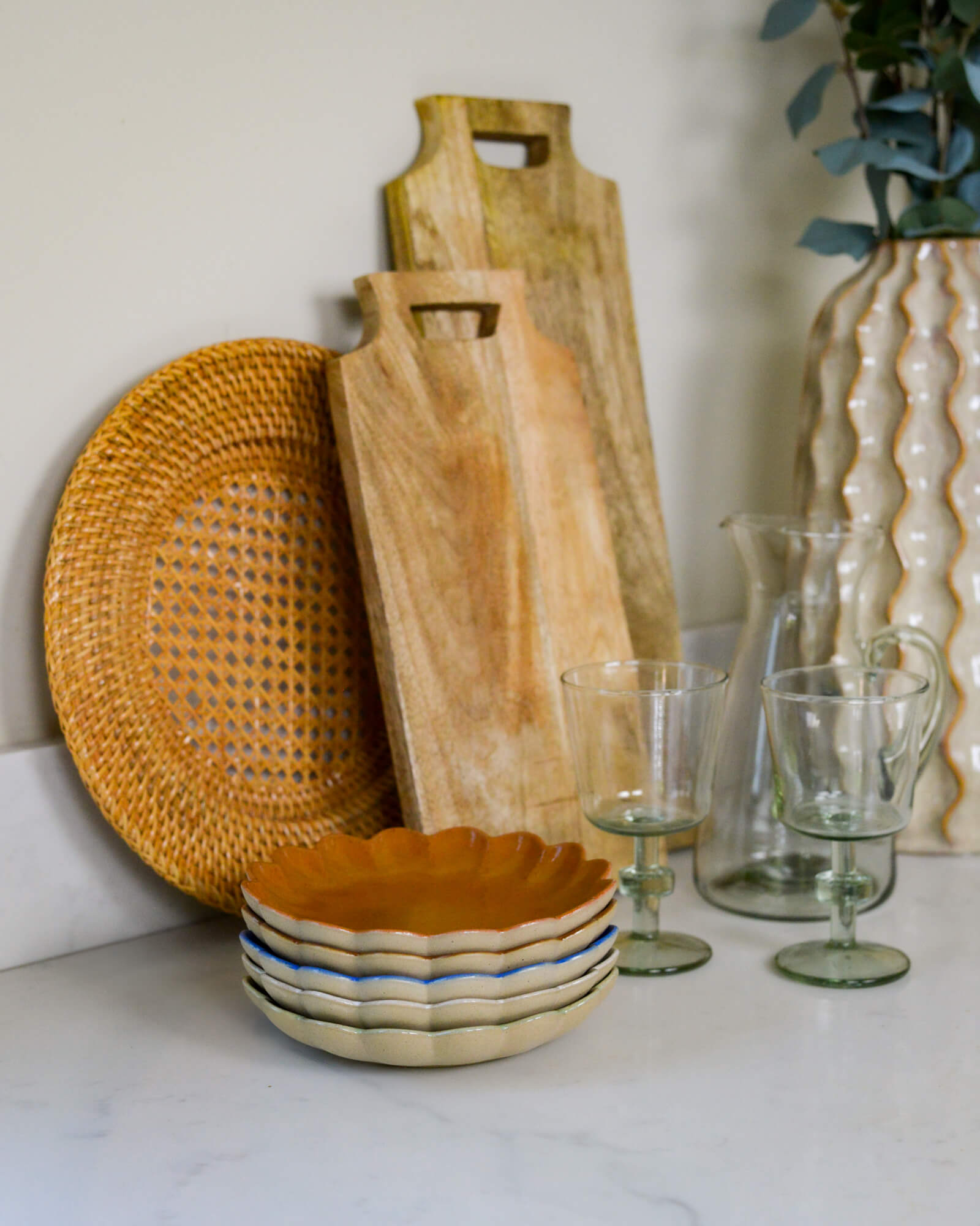 Wooden cutting board, woven plate, ceramic bowl, and glassware on a white surface.