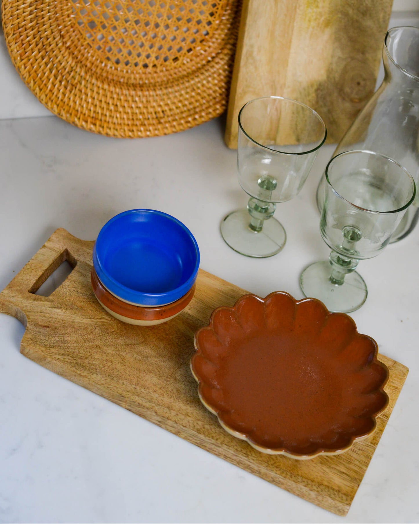 Ceramic bowls and glasses on a wooden grazing paddle with a woven mat in the background.