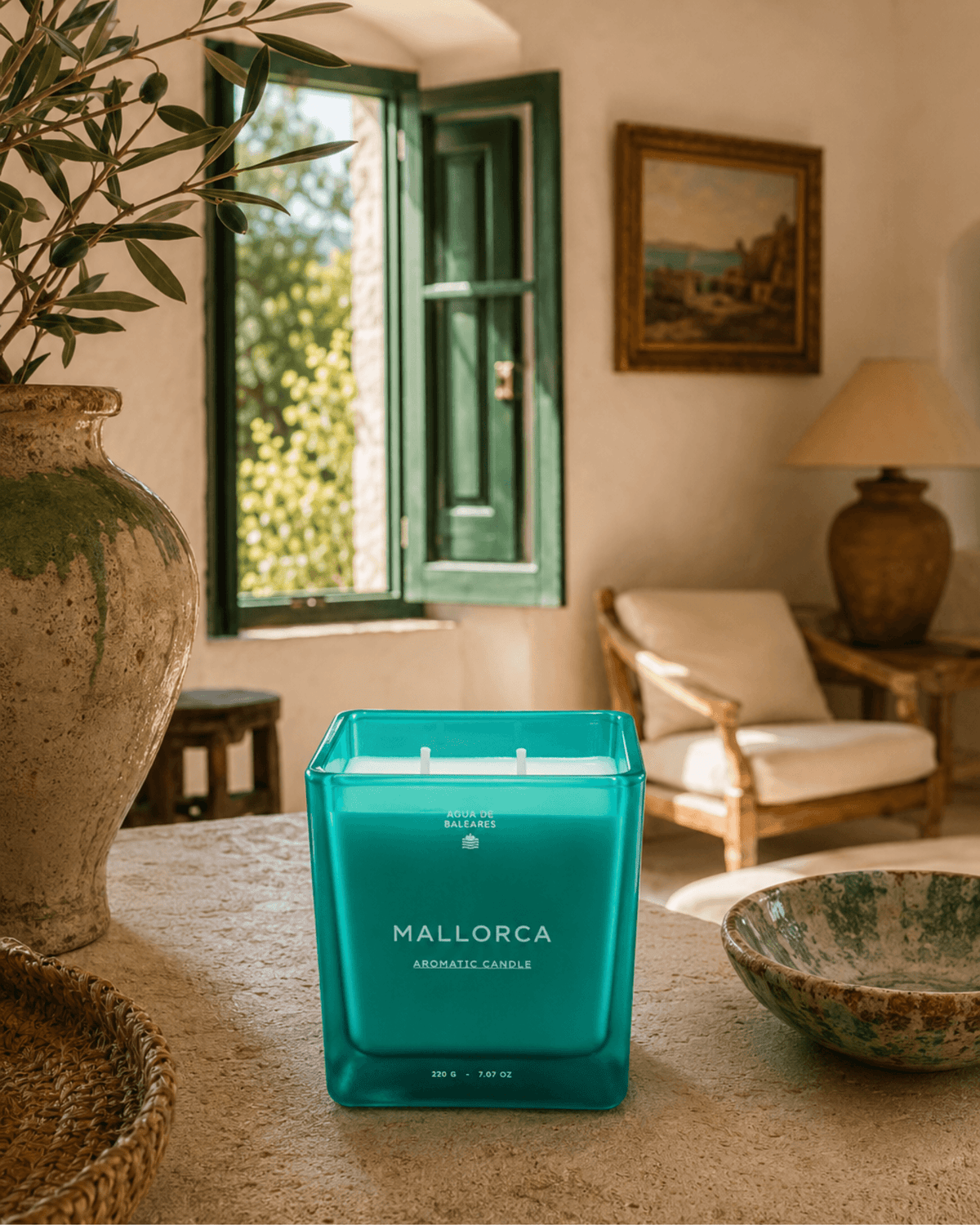 A teal square glass candle labeled 'MALLORCA' sits on a stone table in a warm, rustic interior with a window, chair, lamp, and decorative vases in the background.