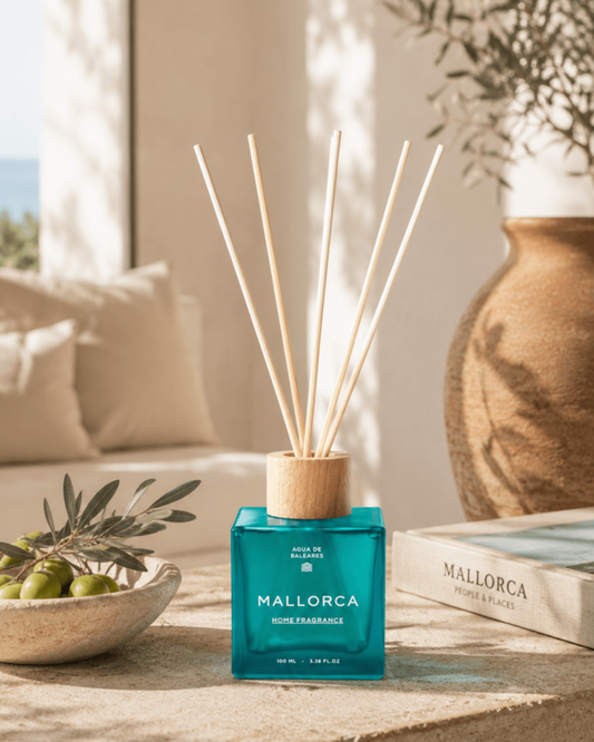A reed diffuser bottle with a translucent teal-blue glass body, a light natural wood collar, and multiple pale reed sticks, labeled MALLORCA, on a rustic wooden mediterranean table