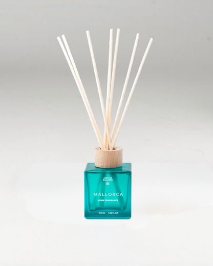 A reed diffuser bottle with a translucent teal-blue glass body, a light natural wood collar, and multiple pale reed sticks, labeled MALLORCA, on a clean light gray background.