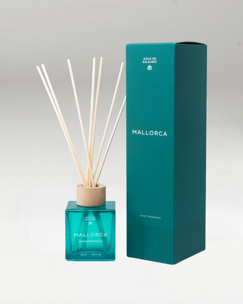 A reed diffuser bottle, and its box, with a translucent teal-blue glass body, a light natural wood collar, and multiple pale reed sticks, labeled MALLORCA, on a clean light gray background.
