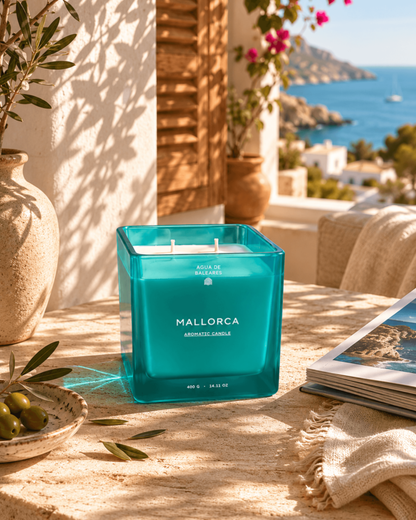 A teal-blue square glass candle labeled 'MALLORCA AROMATIC CANDLE' sits on a sunlit stone table outdoors, with a Mediterranean sea and coastal buildings visible in the background. A vase, olive branch, and open magazine are nearby.