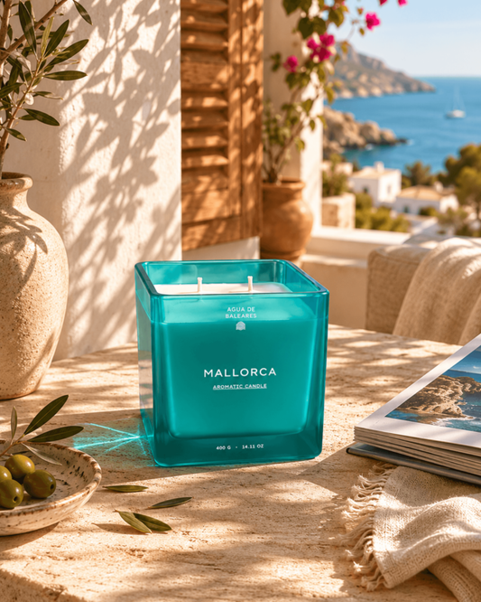 A teal-blue square glass candle labeled 'MALLORCA AROMATIC CANDLE' sits on a sunlit stone table outdoors, with a Mediterranean sea and coastal buildings visible in the background. A vase, olive branch, and open magazine are nearby.