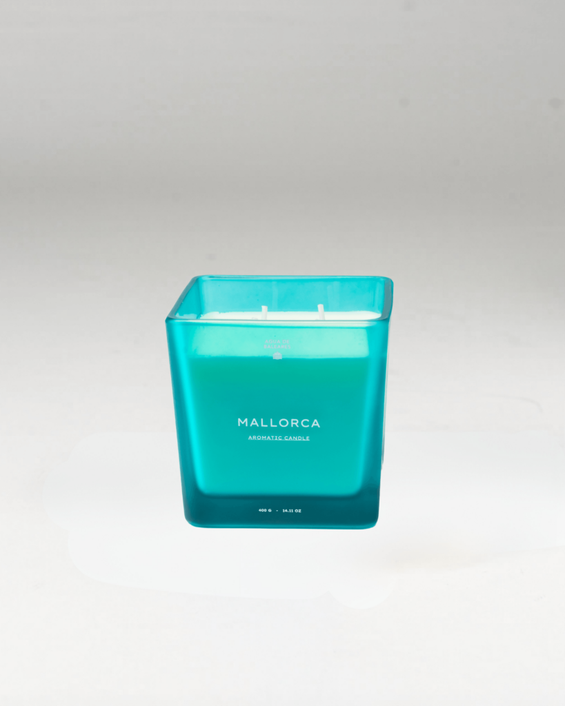 A square glass jar candle with a translucent turquoise blue body and white wax inside, featuring the brand name 'MALLORCA' printed on the front, set against a neutral light gray background.