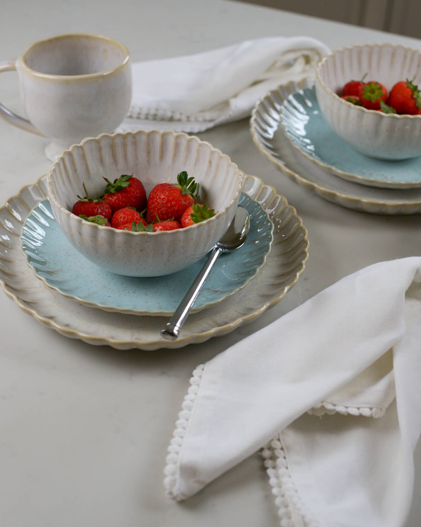 Dining table setting with ceramic scalloped edged plates, cereal bowls, and cups on a light marble surface.