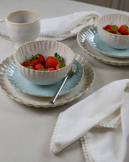 Dining table setting with ceramic scalloped edged plates, cereal bowls, and cups on a light marble surface.