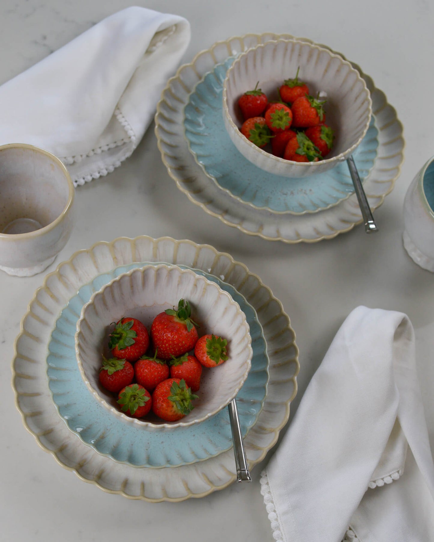 Two ceramic scalloped edge bowls with strawberries on light blue salad plates on a light marble surface with white napkins.
