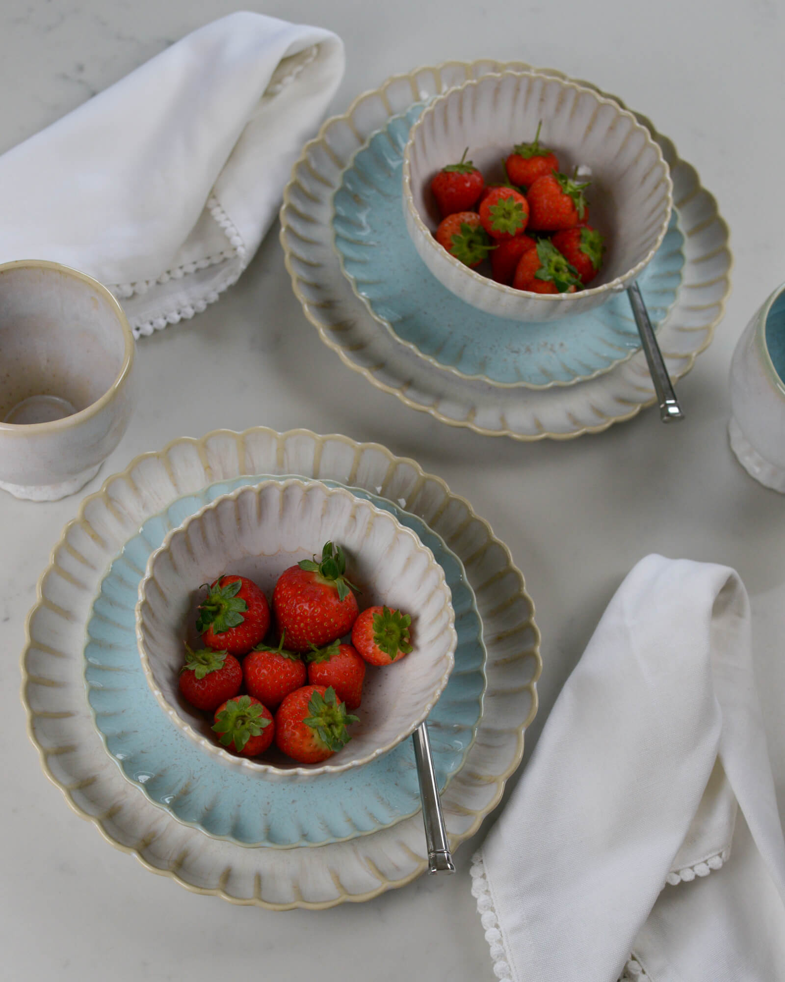 Two ceramic scalloped edge bowls with strawberries on light blue salad plates on a light marble surface with white napkins.