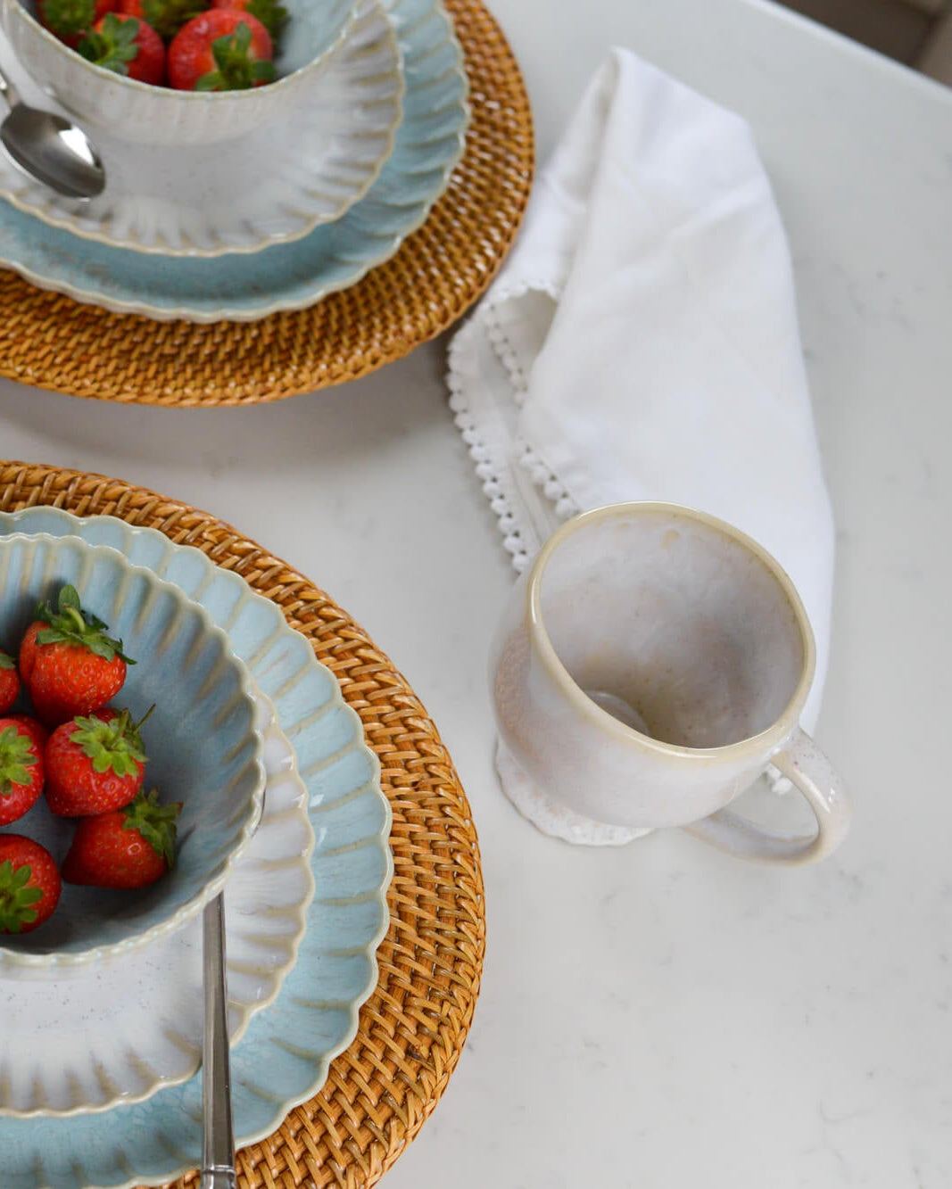 Table setting with ceramic plates, strawberries, a scalloped footed mug, and a white napkin on a light surface.