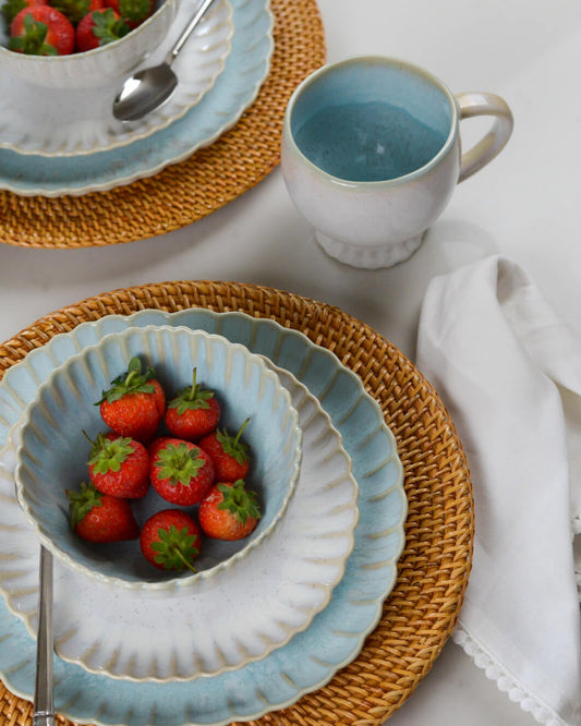 Tea set with large blue ceramic cups and bowls, strawberries, and a woven placemat on a white surface.