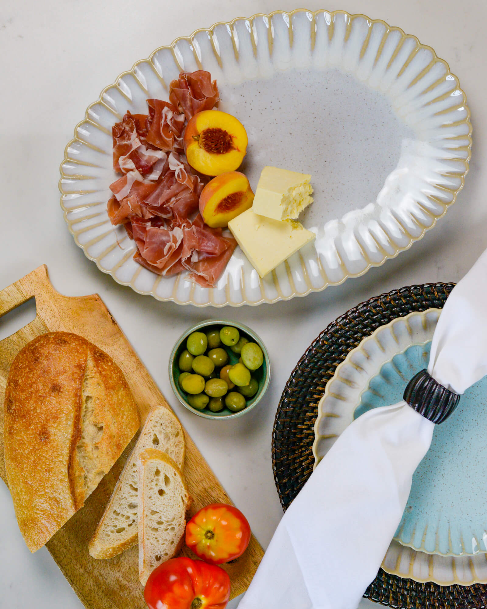 Large oval serving platter with prosciutto, peaches, cheese, bread, and olives on a light marble surface.