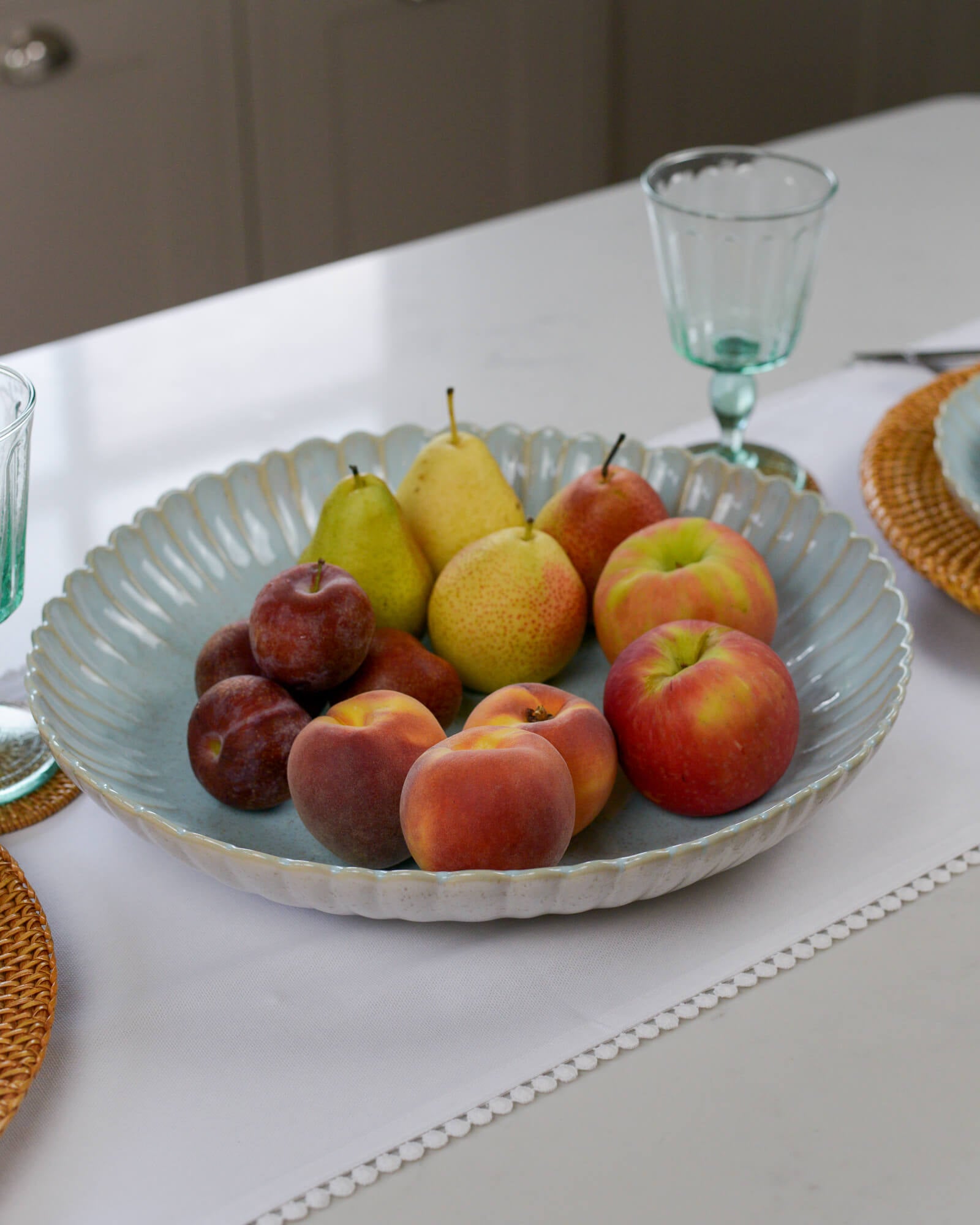 Light blue fruit bowl serving dish with scalloped edge with pears, apples, and plums on a table with glasses and plates.
