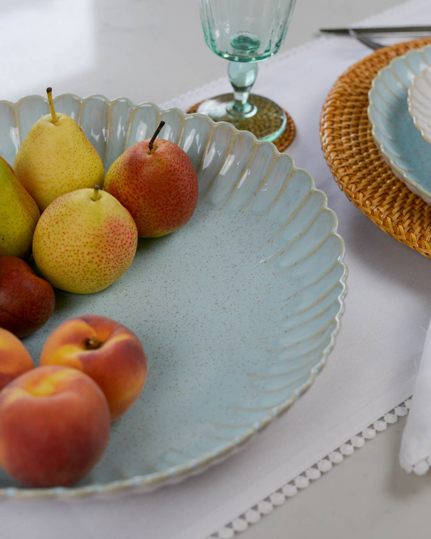 Fruit including pears and peaches in a decorative scalloped edge serving dish with a glass and plates in the background.