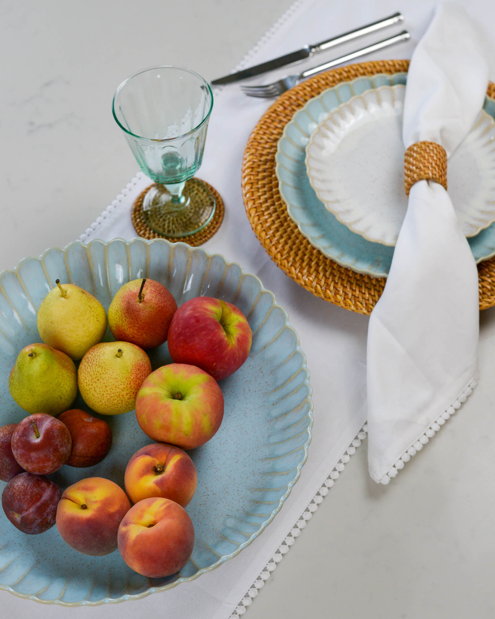 Fruit bowl serving dish with a scalloped edge containing apples, pears, and peaches on a table setting with plates and cutlery.