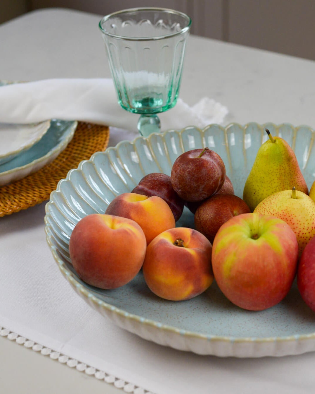 Large fruit bowl serving dish with a scalloped edge containing apples, pears, and plums on a table with glasses and plates.
