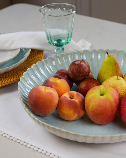 Large fruit bowl serving dish with a scalloped edge containing apples, pears, and plums on a table with glasses and plates.