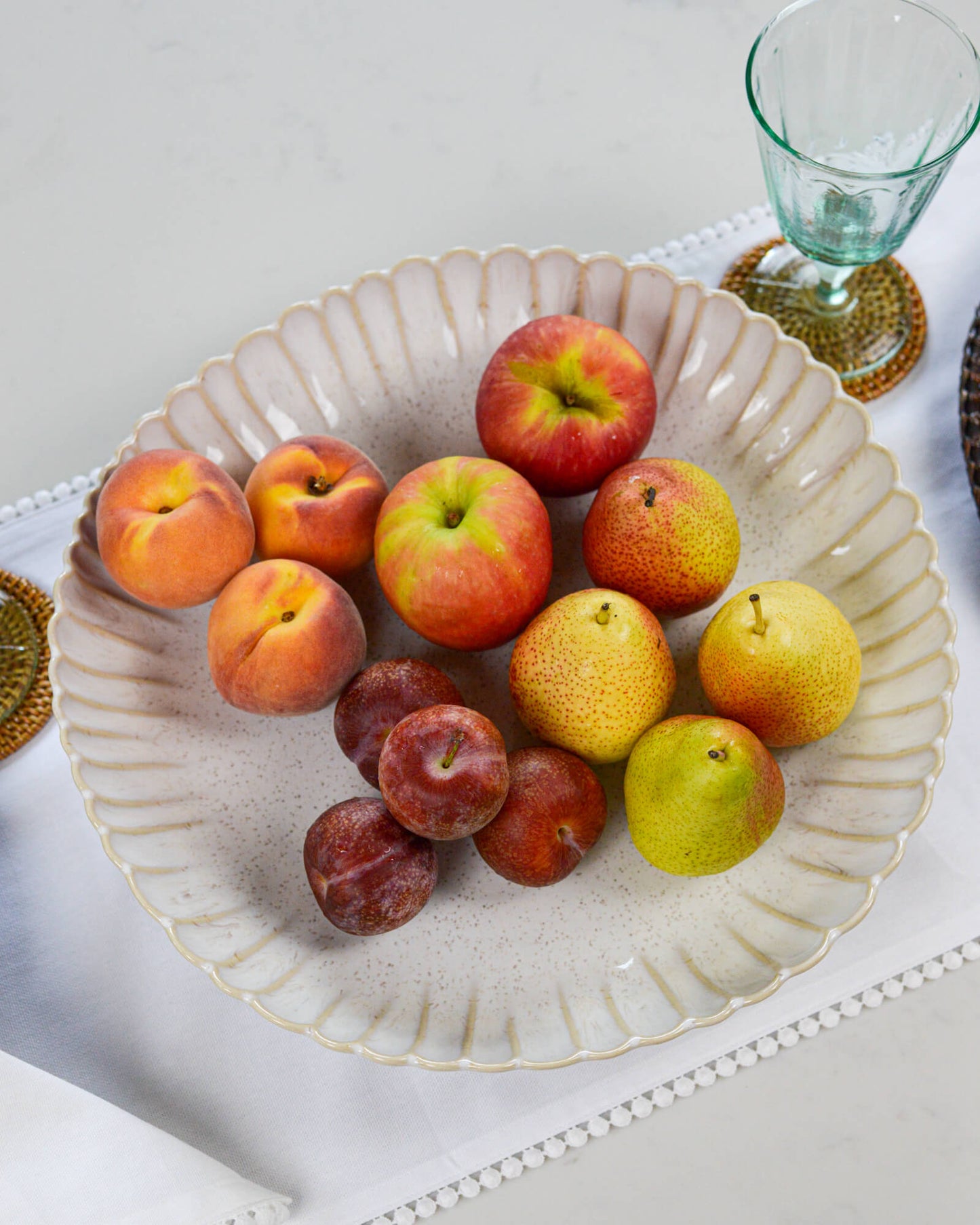 Fruit including apples, peaches, and pears in a decorative serving dish with a glass and coaster in the background.