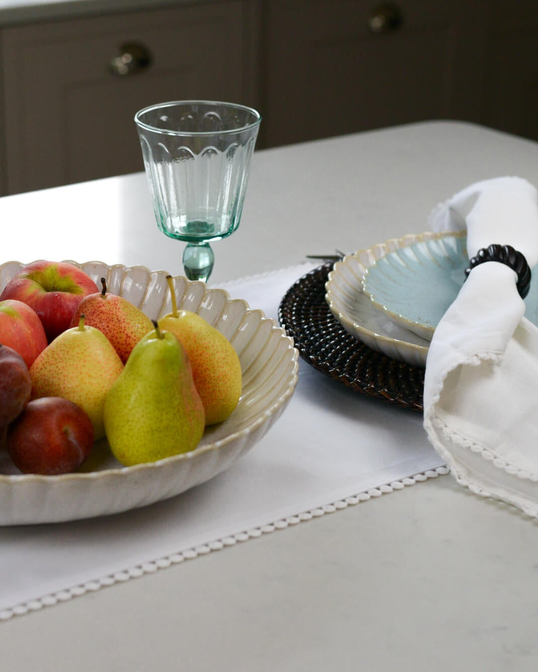 Fruit bowl with scalloped edge containing pears and apples next to a glass on a table.