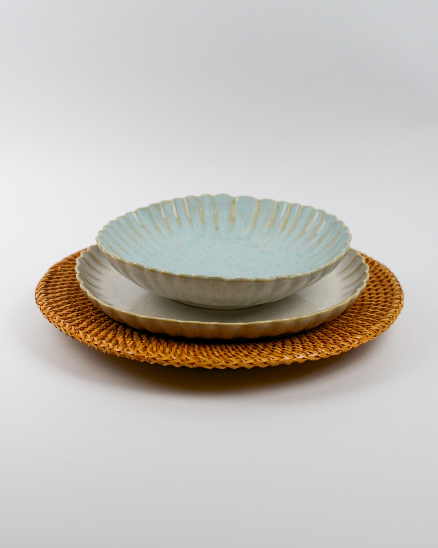 Set of ceramic scalloped edge pasta bowl and matching plates on a woven rattan charger plate with a white background