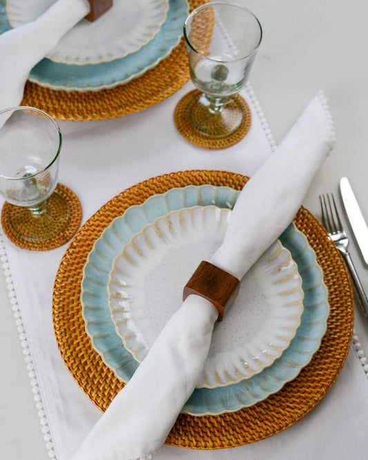 Dining table setting with white and light blue ceramic, stoneware plates, woven placemats, and cutlery on a white table runner.