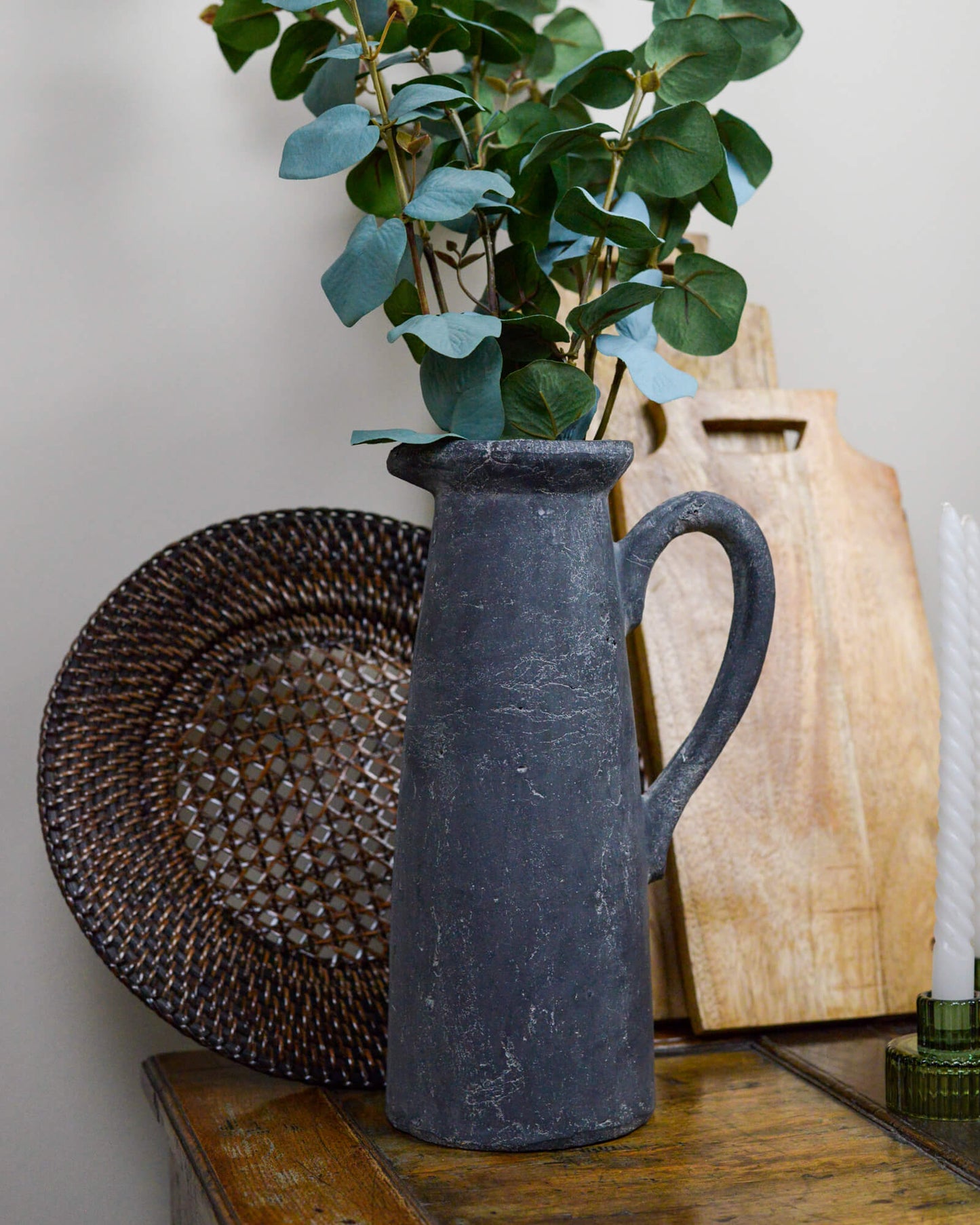 Dark gray pitcher with green leaves on a wooden surface with wooden chopping boards and rattan charger plate 