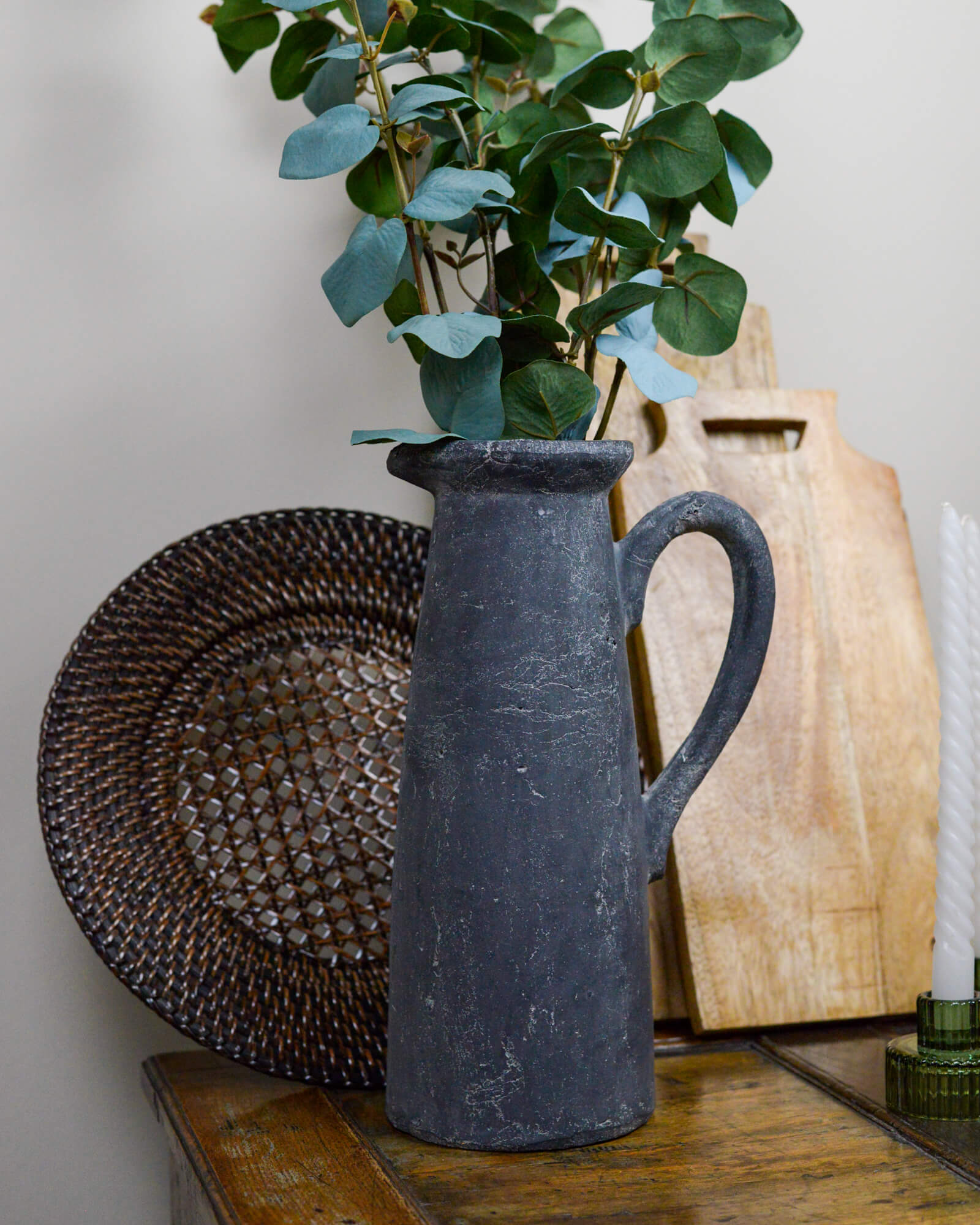 Dark gray pitcher with green leaves on a wooden surface with wooden chopping boards and rattan charger plate 