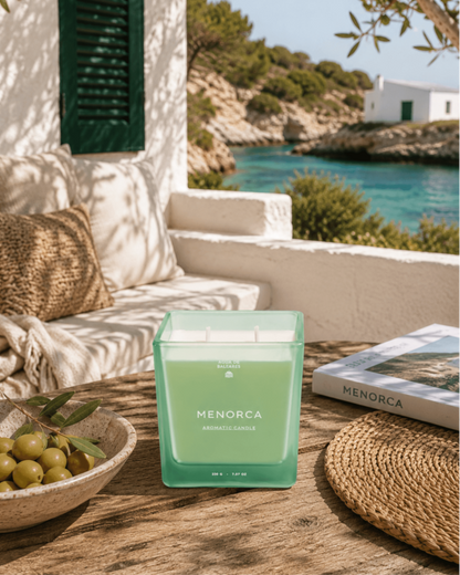 A square glass jar candle in a mint green  sits on a rustic wooden table in a sunlit outdoor setting with a Mediterranean-style white wall, turquoise water, olive branches, and a book labeled MENORCA.