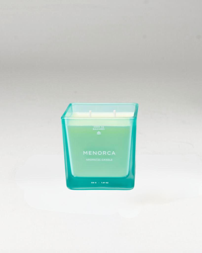 A square glass jar candle with a translucent turquoise-green gradient design and white wax inside, labeled 'MENORCA' on the front, set against a neutral light gray background.