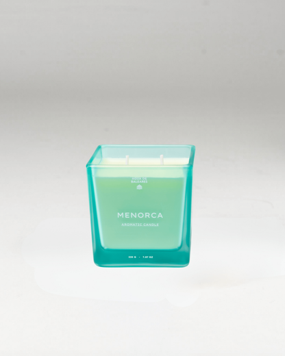 A square glass jar candle with a translucent turquoise-green gradient design and white wax inside, labeled 'MENORCA' on the front, set against a neutral light gray background.