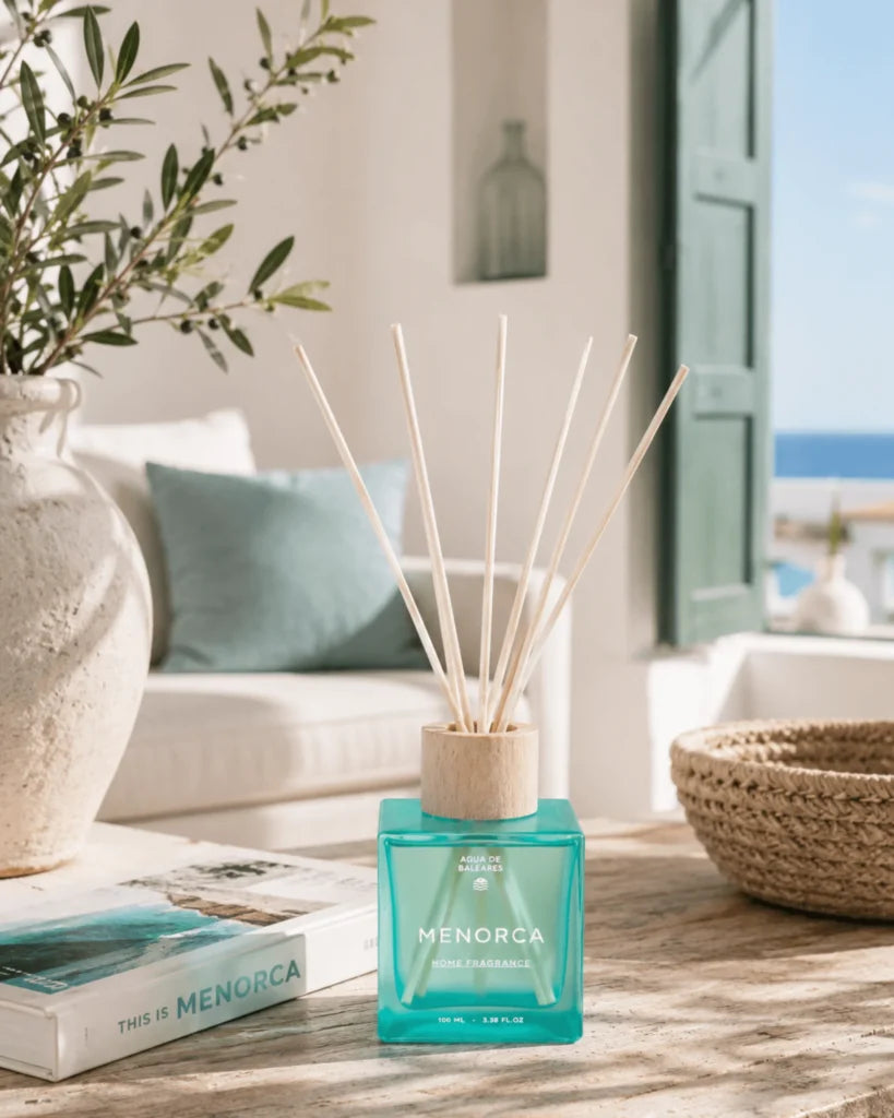 A reed diffuser bottle with a translucent turquoise blue glass body and natural wood reeds, placed on a sunlit wooden table in a bright, modern interior setting with a sofa, vase, basket, and window view.
