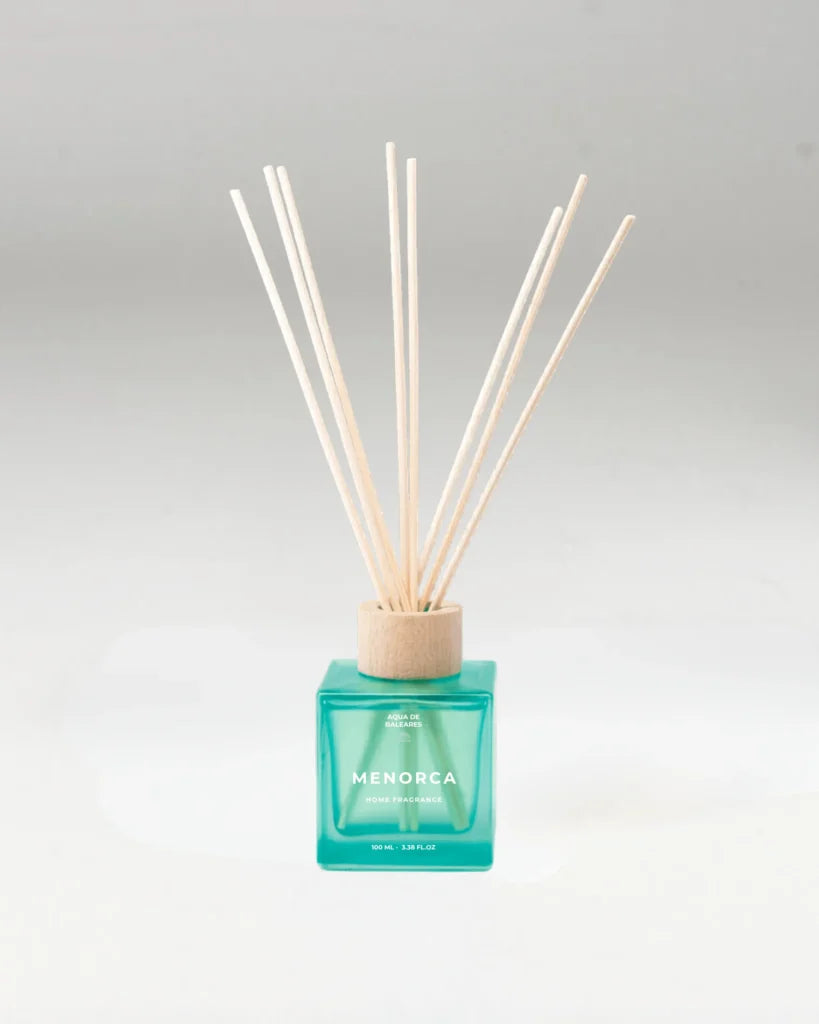 A reed diffuser with a translucent turquoise glass bottle and natural wood reeds, labeled 'MENORCA' on the front, set against a neutral light background.