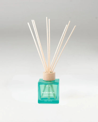A reed diffuser with a translucent turquoise glass bottle and natural wood reeds, labeled 'MENORCA' on the front, set against a neutral light background.