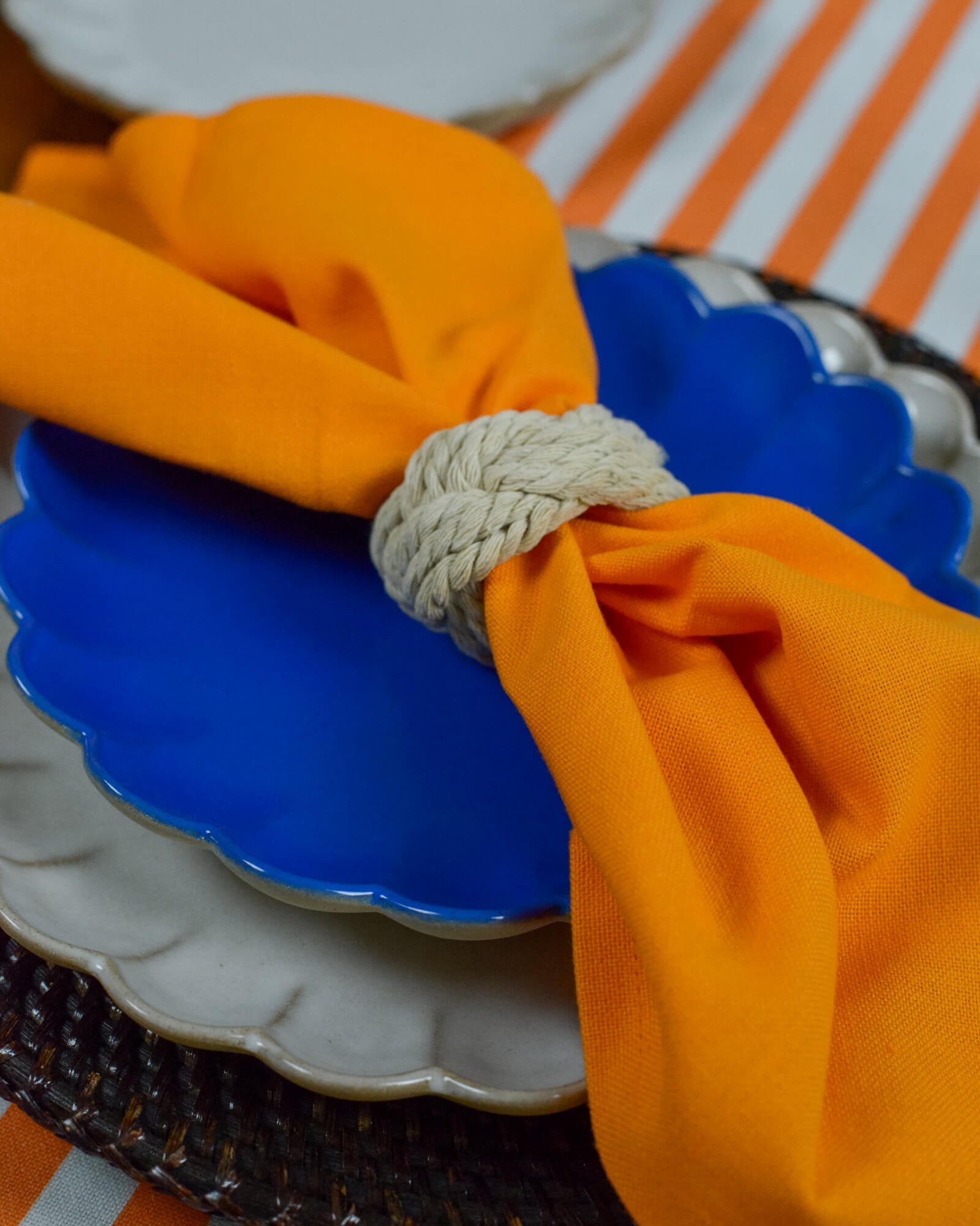 Orange napkin held with a rope napkin ring on a blue scalloped side plate with a striped background