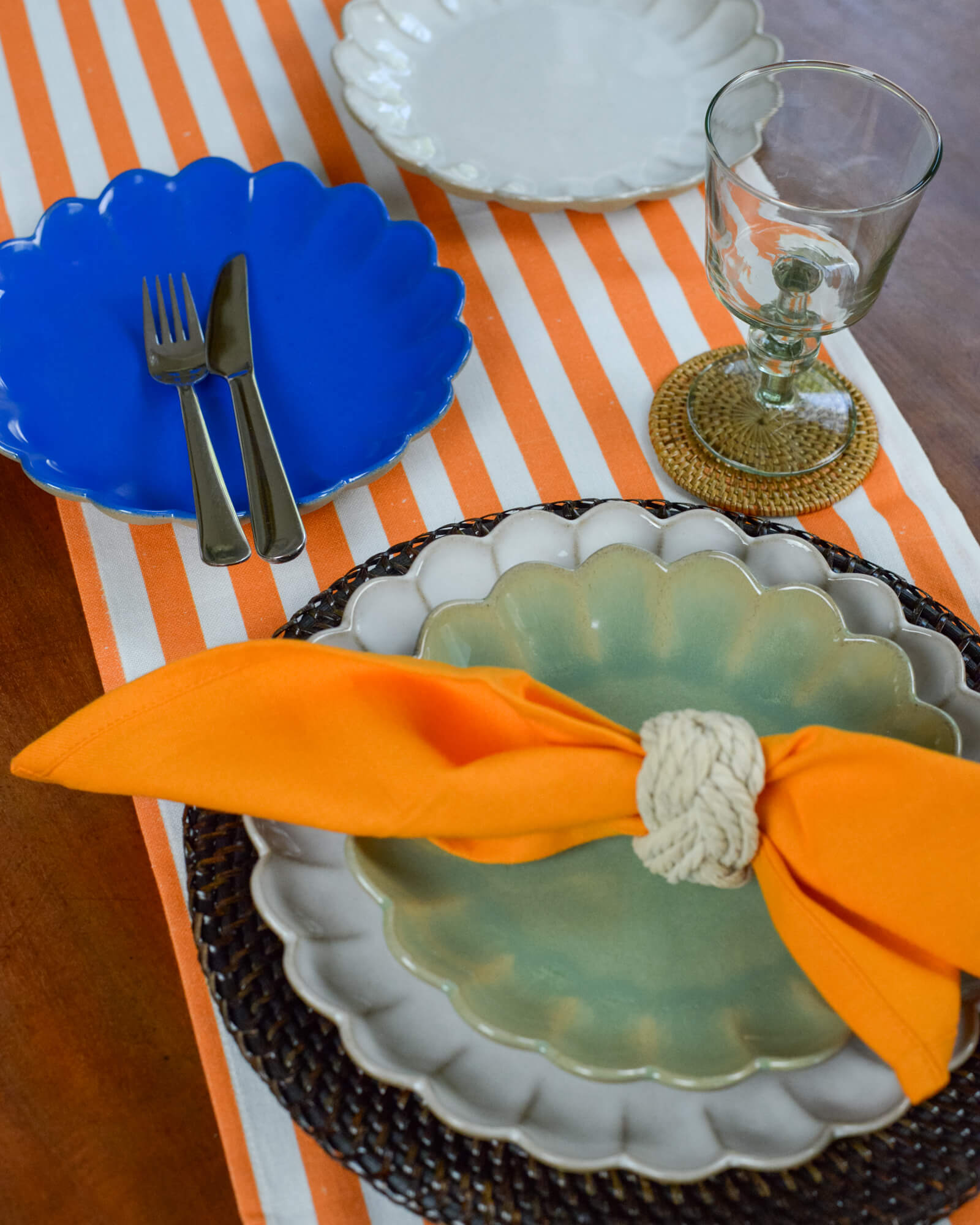 Table setting with blue plate, silverware, orange napkin, and glass on a striped orange cotton table runner.