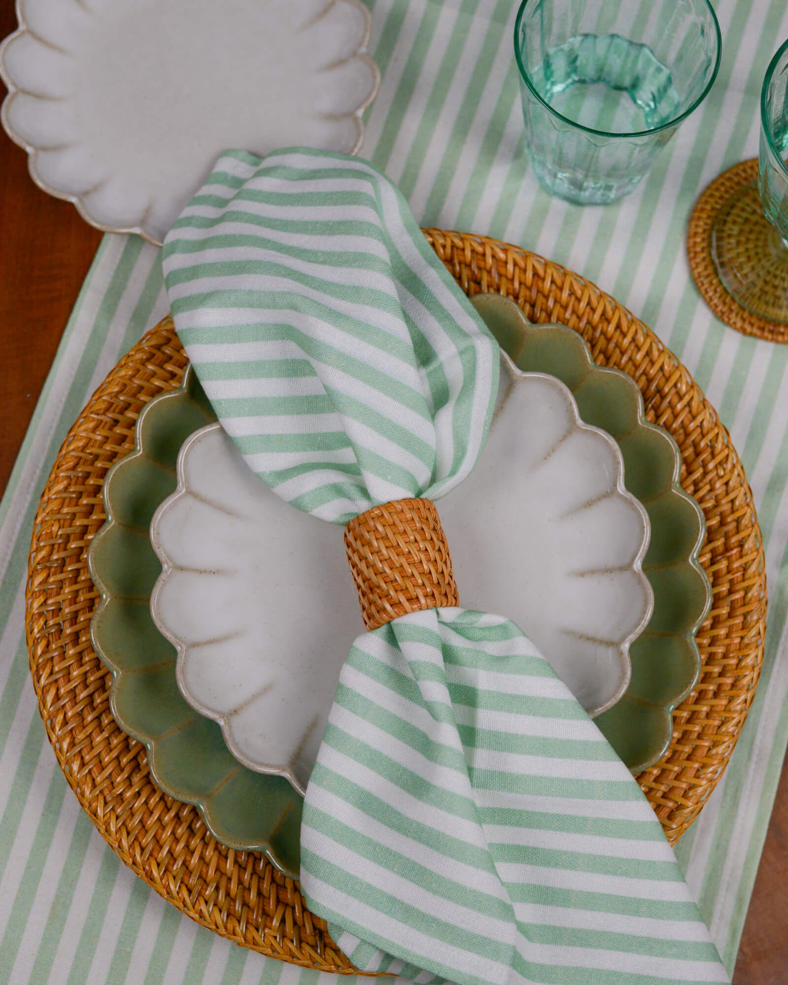 Table setting with a shell-shaped plate on a rattan charger plate, green and white striped napkin, and clear glasses on a striped tablecloth.