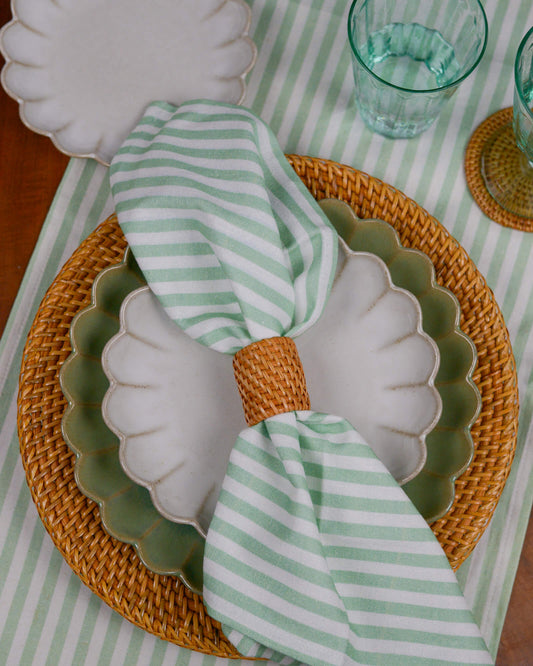 Table setting with a shell-shaped plate on a rattan charger plate, green and white striped napkin, and clear glasses on a striped tablecloth.