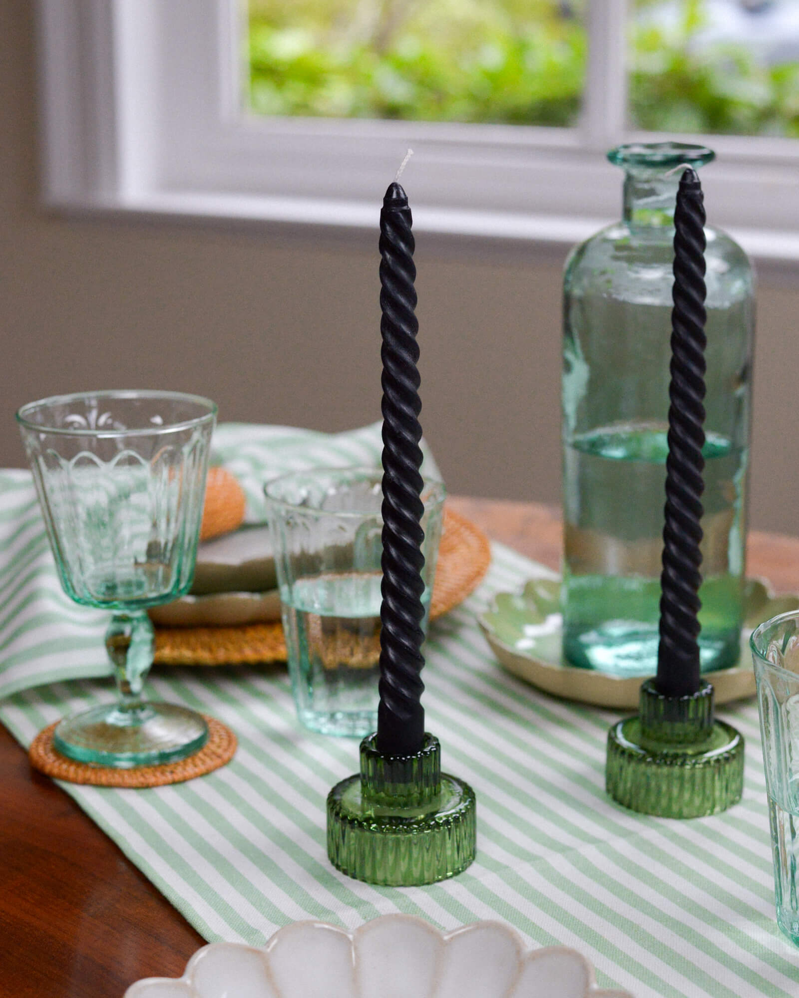 Black twisted candles on green candle holders on a table with glasses and a striped tablecloth.