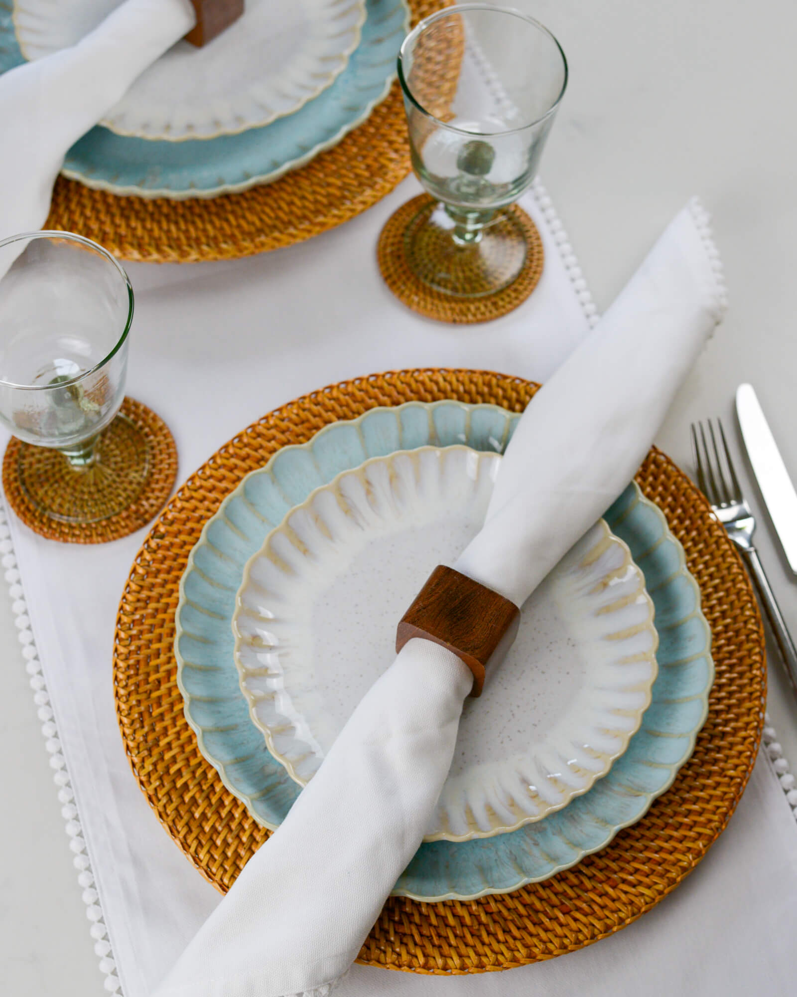 Dining table setting with ceramic plates, white cotton napkins, woven placemats, and cutlery on a white cotton table runner.
