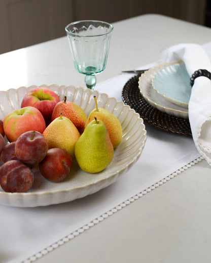 Fruit bowl with apples, pears, and plums on a table with a white pearl edged table runner, glass and plates.