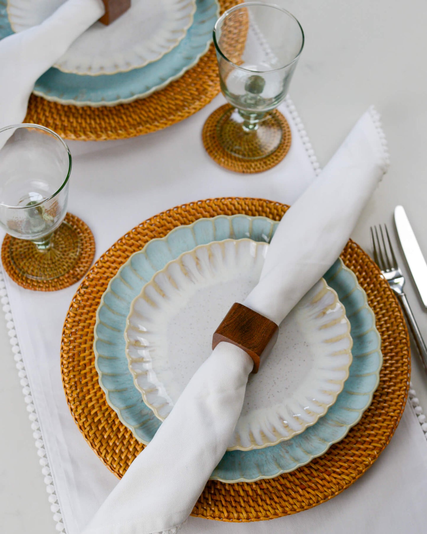 Dining table setting with ceramic plates, woven placemats, and cutlery on a white tablecloth.