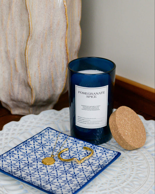 Candle labeled 'Pomegranate Spice' on a decorative plate with a textured vase in the background.