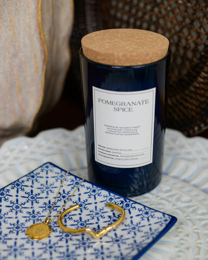Candle labeled 'Pomegranate Spice' with cork lid on a textured surface with jewelry.
