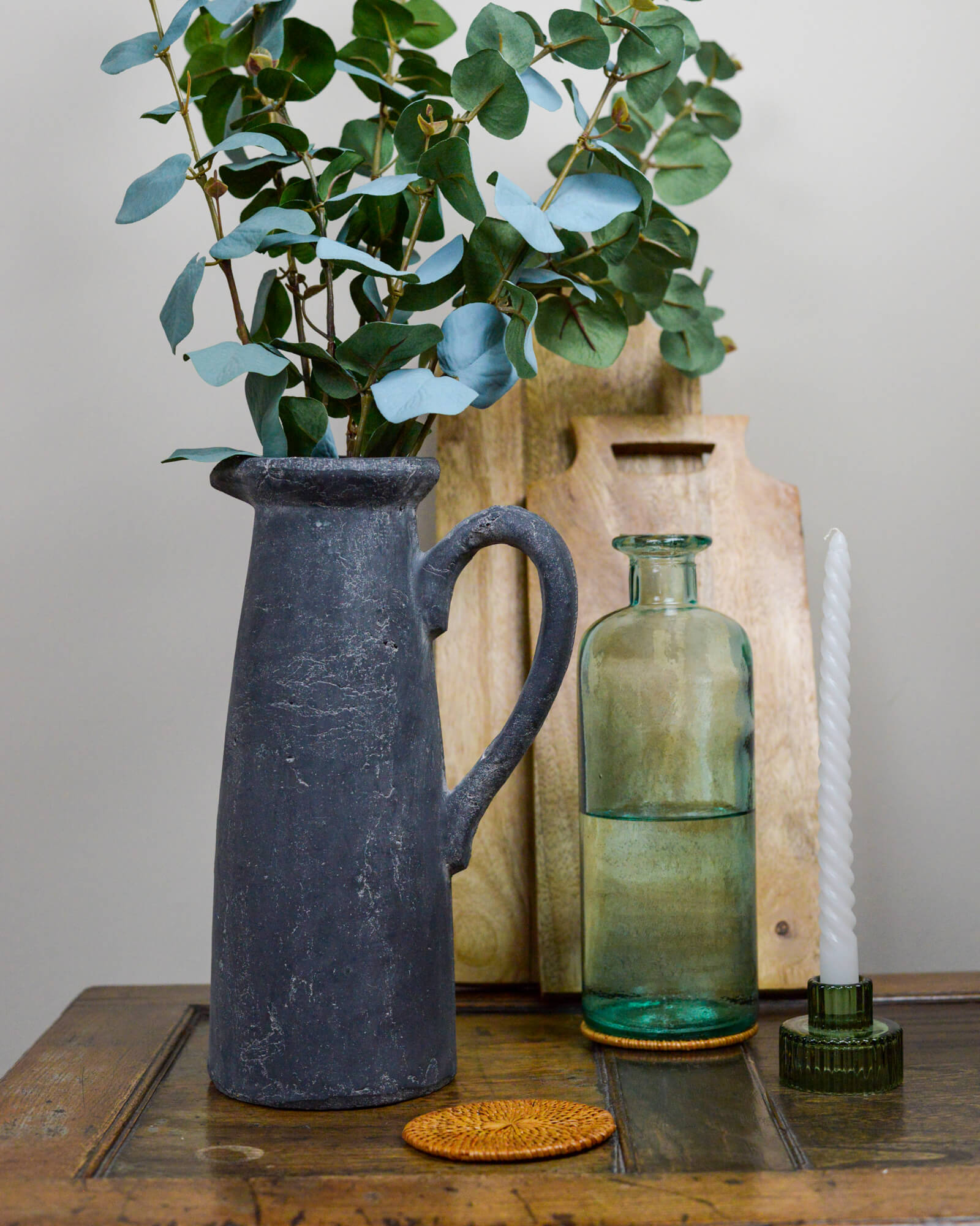 Decorative still life with a pitcher, green bottle on a rattan coaster, and candle on a wooden surface.
