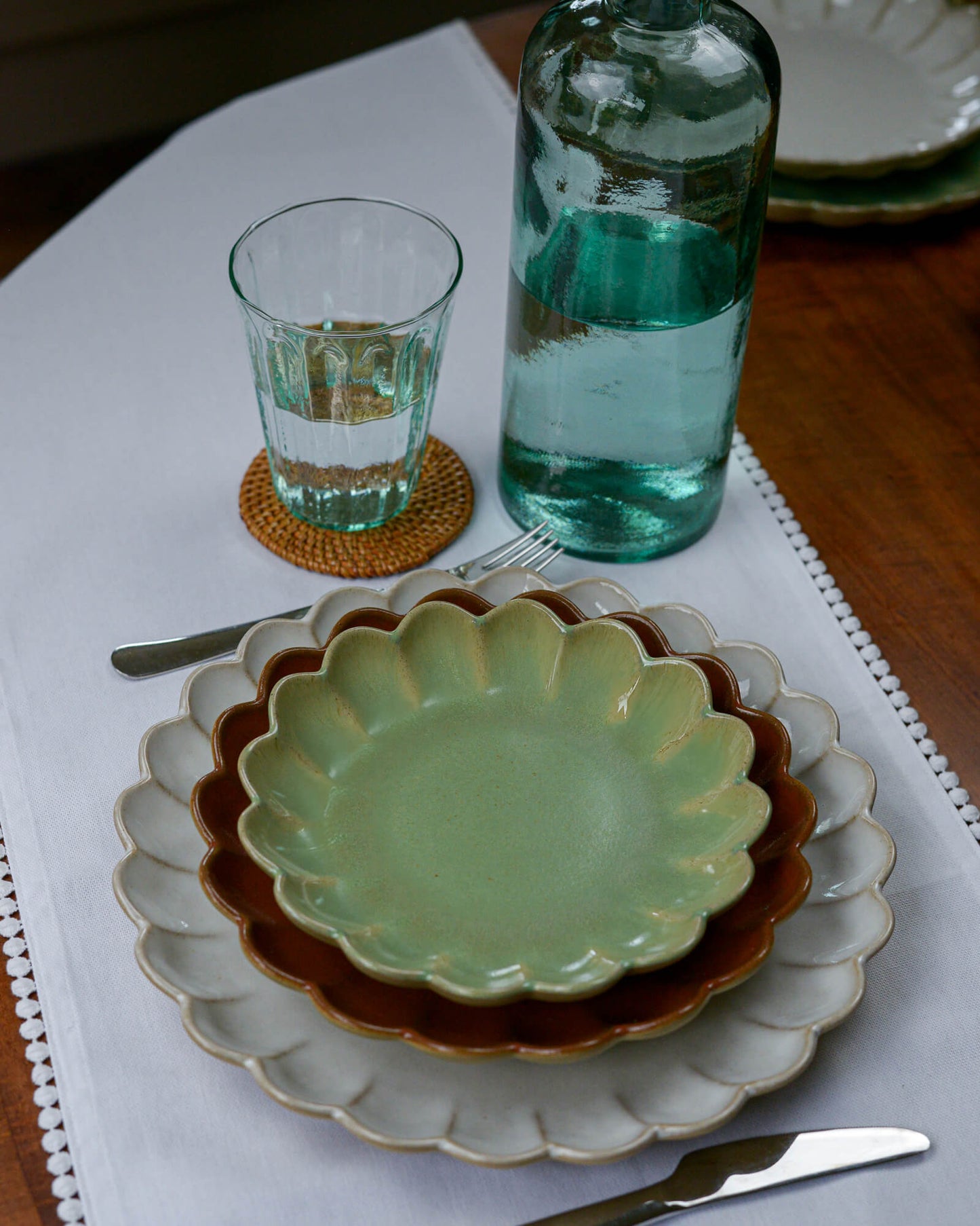 Set table with green ceramic plates, glass, and bottle on a white tablecloth with rattan coasters.