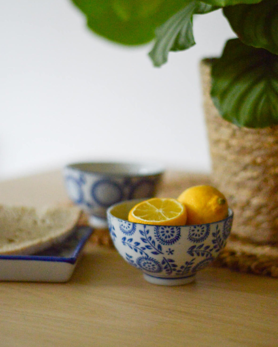 Blue and white ceramic bowl with lemons on a wooden surface, with a plant in the background.