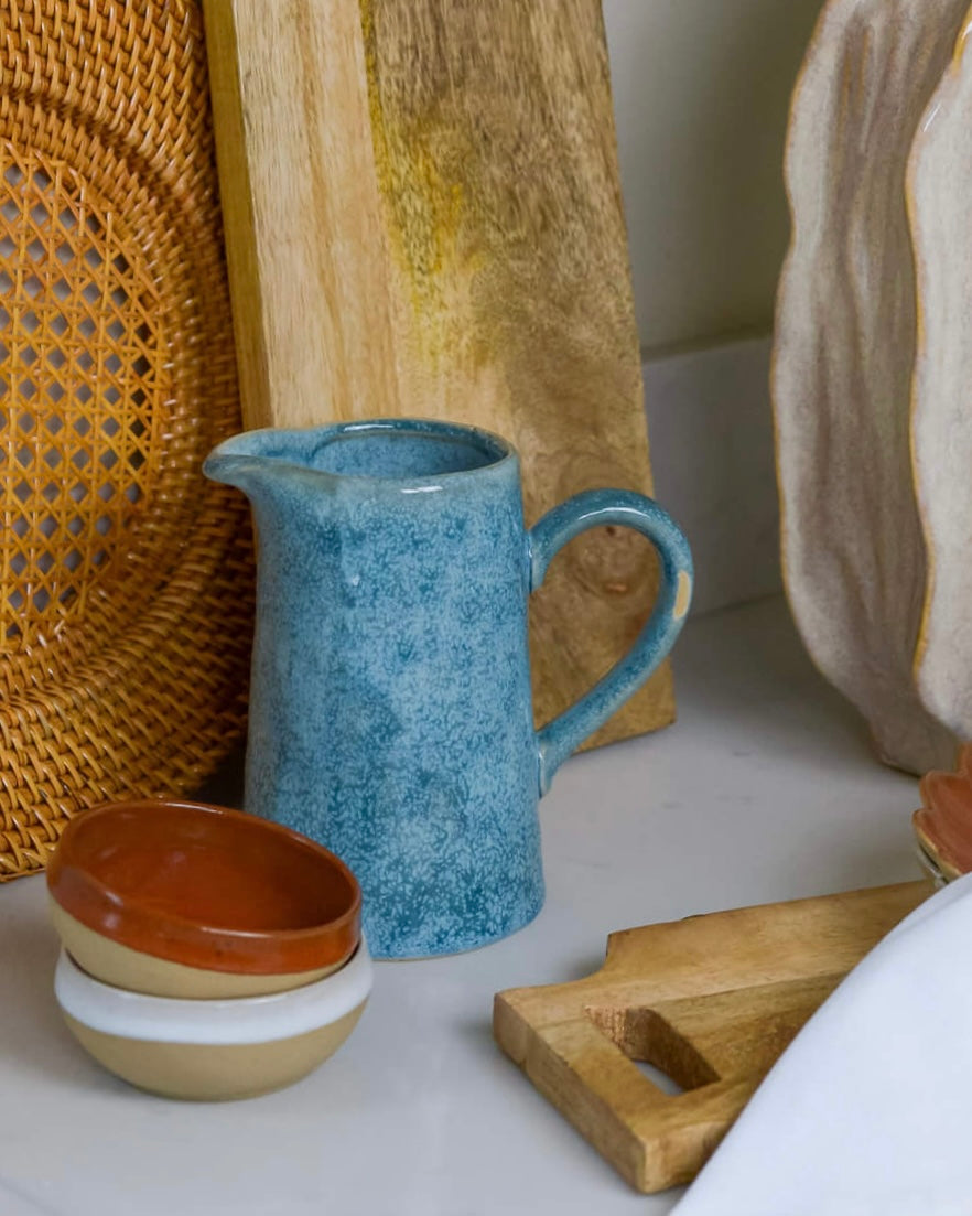 Set of ceramic items including a blue pitcher, brown and white dipping bowls, and a wooden cutting board on a white marble surface.