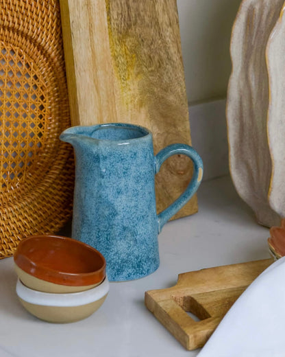 Set of ceramic items including a blue pitcher, brown and white dipping bowls, and a wooden cutting board on a white marble surface.